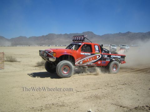1990 Ford Bronco - orange Maxxis-sponsored off-road race truck numbered 785 kicks up dust across a flat desert course.