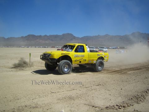 1990 Ford Bronco - yellow off-road race truck numbered 719 speeds across a flat desert race course backed by distant mountains.