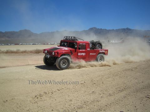 1990 Ford Bronco - red Hummer-style off-road race truck slides through a dusty desert turn with mountains behind.