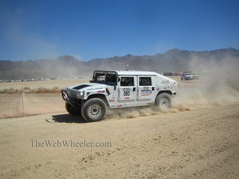 1990 Ford Bronco - white Hummer-style off-road race truck numbered 860 races across a dusty desert course near mountains.
