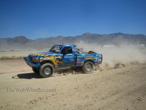 1990 Ford Bronco - blue-and-yellow flame-liveried off-road race truck numbered 013 kicks up dust on a flat desert course.