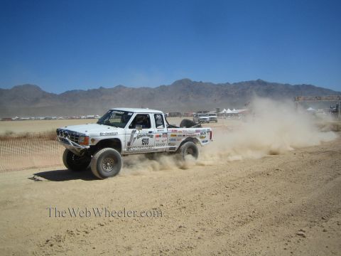 1990 Ford Bronco - white off-road race truck numbered 900 throws up dust as it races across a flat desert course near mountains.