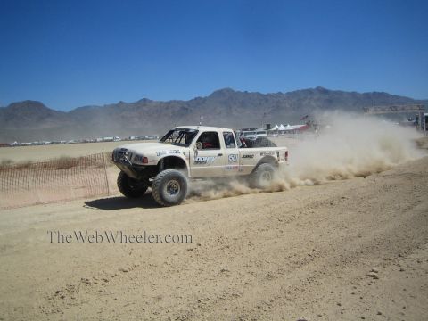 1990 Ford Bronco - white off-road race truck sprays dust as it races along a graded desert track with mountains behind.