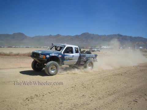 1990 Ford Bronco - white-and-blue off-road race truck kicks up dust along a graded desert race track backed by mountains.