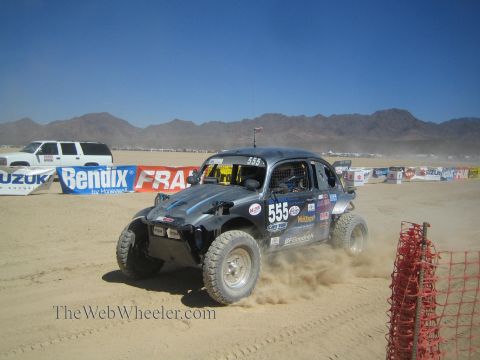 1990 Ford Bronco - black VW Beetle-based Baja Bug numbered 555 sits on a desert race course near sponsor banners.