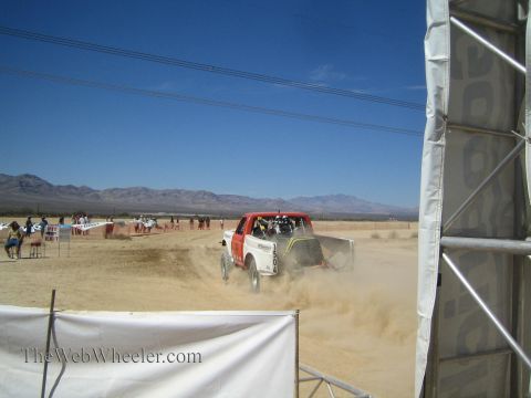 1990 Ford Bronco - off-road race truck speeds away through dust past a banner-framed staging area in the desert.
