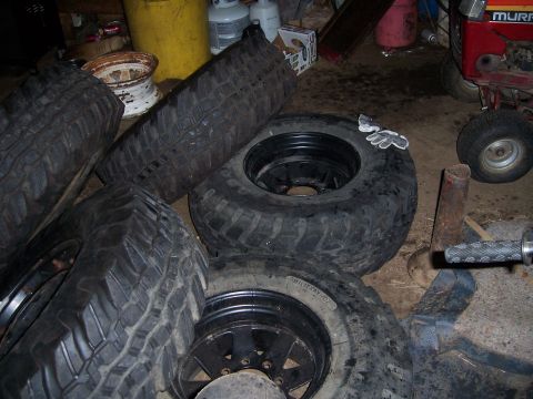 1983 Ford Bronco - Several off-road tires and black rims are piled in a garage, with a rusty white rim in the background.