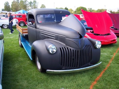1972 Ford Ranchero - Matte black 1940s pickup truck with hood open at a car show, with red sports cars in the background.