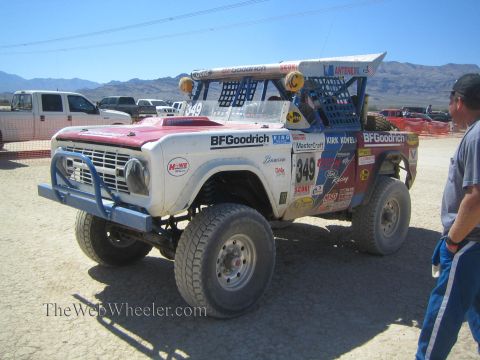 1990 Ford Bronco - red-and-white early Ford Bronco race truck numbered 349 is parked on a dirt lot as a man stands beside it.