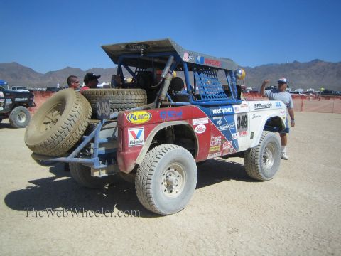 1990 Ford Bronco - red-and-white early Ford Bronco race truck numbered 349 with rear spare tires sits in the desert as crew members stand around it.