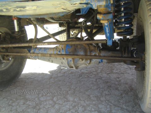 1990 Ford Bronco - close-up underbody shot showing a vehicle's blue-painted front axle, leaf springs, and coil shock over hard desert ground.
