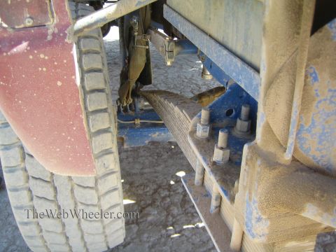 1990 Ford Bronco - Close-up of an off-road truck's rear suspension showing a blue-painted leaf spring pack, U-bolts and a dirty mud flap behind a worn tire.