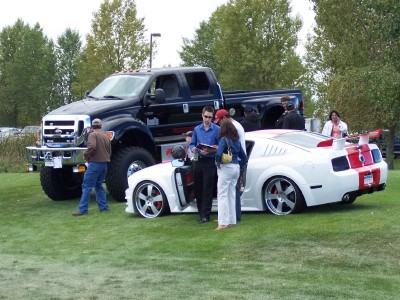 1972 Ford Ranchero - Two vehicles, a white sports car and a large black truck, are parked on a grassy field with people around them.