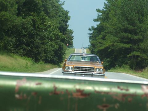 1966 Ford Ranchero - A gold sedan is seen driving down a tree-lined road, with another vehicle in the distance.