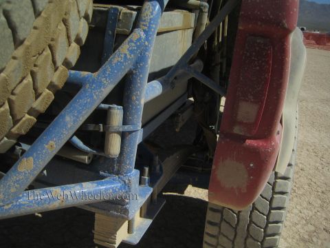 1990 Ford Bronco - Rear undercarriage of a desert off-road truck showing a dust-coated blue tubular frame, leaf spring and a faded red bumper.