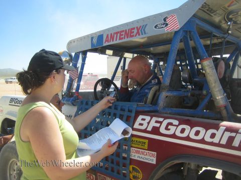 1990 Ford Bronco - woman in a yellow top holds a clipboard beside a blue off-road race truck with Antenex and Ford decals while the seated driver gestures.