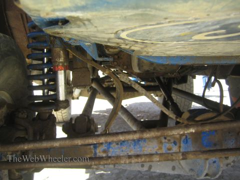 1990 Ford Bronco - Underbody view of a blue off-road truck revealing a coilover shock, suspension links and a battered painted frame rail.