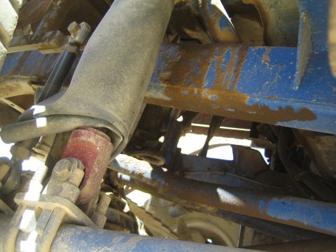 1990 Ford Bronco - Detail of a worn shock absorber with a red collar amid rusty and blue-painted tubular suspension links under a vehicle.