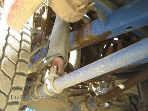 1990 Ford Bronco - Underbody shot of a blue tube-frame off-road truck showing a shock with red collar, a heavy link bar and a tire at left.