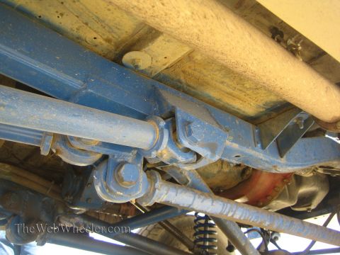 1990 Ford Bronco - Close underbody view of a blue off-road truck with driveshaft, U-joint, steering linkage and a glimpse of a coil spring.