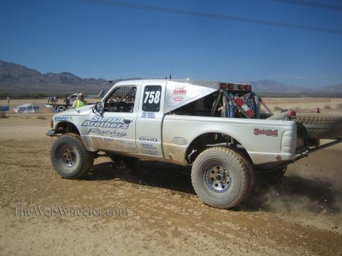 1990 Ford Bronco - white Sletten Brothers Racing number 758 off-road truck carrying a spare tire drives across a dusty desert flat.
