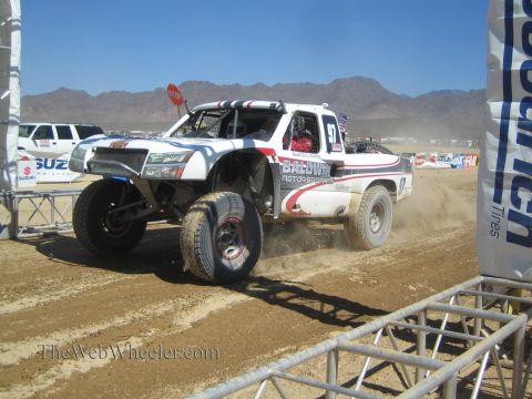 1990 Ford Bronco - white and silver Baldwin Motorsports off-road race truck launches off a bump, lifting a front wheel near a Suzuki banner.