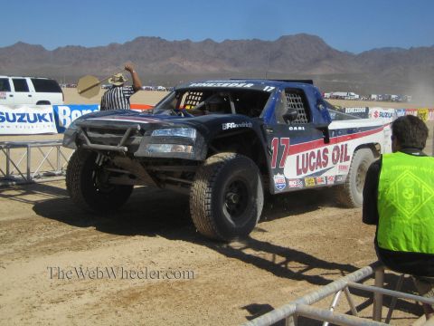 1990 Ford Bronco - black Lucas Oil number 17 off-road race truck rounds a dirt turn as a flagger signals and a green-vested official watches.