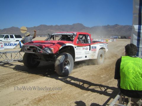 1990 Ford Bronco - red and white number 4 off-road race truck corners on a dusty course as a flag marshal and seated official look on.