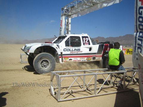 1990 Ford Bronco - white Collins Motorsports number 12 off-road race truck speeds beneath a BFGoodrich start arch as an official kneels nearby.