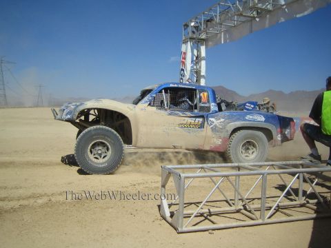 1990 Ford Bronco - mud-caked blue off-road race truck slides sideways through dust beneath a metal start arch on a desert course.