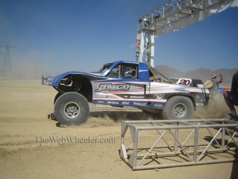1990 Ford Bronco - blue and white Jimco-built number 20 off-road race truck throws dust as it powers through a turn past a flag marshal.