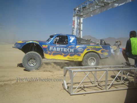 1990 Ford Bronco - blue Fabtech number 7 off-road race truck with yellow flames and Toyo Tires livery races through dust past a kneeling official.