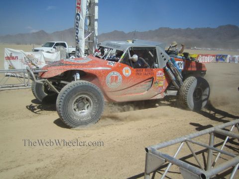 1990 Ford Bronco - orange tube-frame single-seat off-road race buggy numbered 401 corners on a dusty desert course past banners.