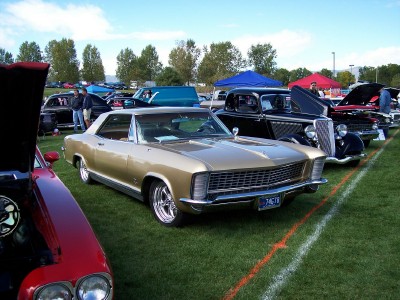1972 Ford Ranchero - A gold 1965 Buick Riviera is displayed at an outdoor car show alongside other classic vehicles.