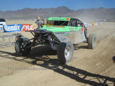 1990 Ford Bronco - green Goodyear-sponsored number 110 open tube-frame off-road buggy drives on a dusty course as a flagger raises a flag.