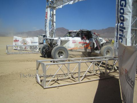1990 Ford Bronco - white open-frame off-road race buggy speeds through heavy dust beneath a BFGoodrich start arch behind safety barriers.