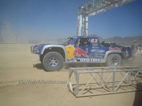 1990 Ford Bronco - blue Red Bull number 83 off-road race truck with Toyo Tires and McMillin Homes decals races beneath a metal start arch.