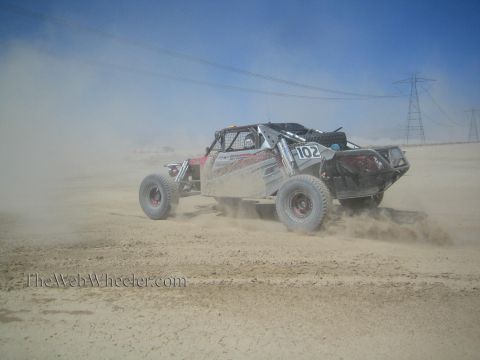 1990 Ford Bronco - silver and red number 102 open-frame off-road race buggy charges through a thick dust cloud across a desert flat.