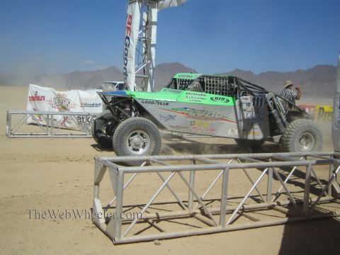1990 Ford Bronco - green and white tube-chassis off-road race buggy launches from a desert start line beneath a BFGoodrich banner arch amid dust.