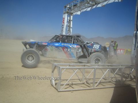 1990 Ford Bronco - blue tube-chassis off-road race buggy numbered 115 races through dust beneath a BFGoodrich banner arch in the desert.