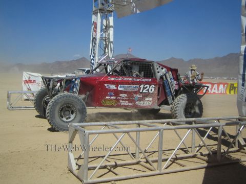 1990 Ford Bronco - dark red off-road race buggy numbered 126 races from a desert start line beneath a BFGoodrich banner arch in dust.