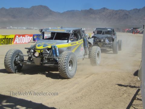 1990 Ford Bronco - yellow and gray off-road race buggy numbered 118 leads a darker buggy along a sandy desert course lined with banners.
