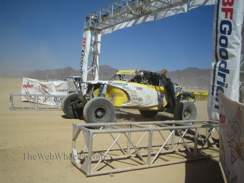 1990 Ford Bronco - yellow and white off-road race buggy passes beneath a BFGoodrich banner start arch on a dusty desert course.