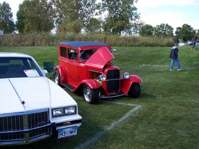 1972 Ford Ranchero - A vintage red hot rod sedan is displayed at an outdoor car show, with its hood open, alongside another vehicle.