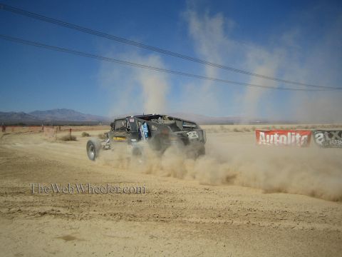 1990 Ford Bronco - off-road race buggy speeds away on a wide dusty desert course past advertising banners and power lines.