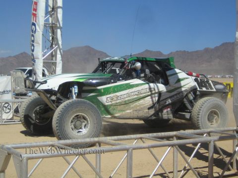 1990 Ford Bronco - green and white off-road race buggy with construction-company livery races across a dusty desert course.