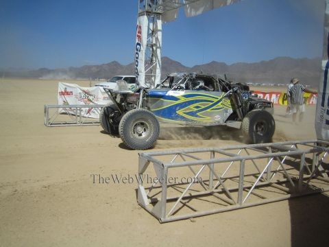 1990 Ford Bronco - blue and yellow off-road race buggy launches from a desert start line beneath a BFGoodrich banner arch in dust.