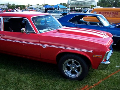 1972 Ford Ranchero - Red muscle car with racing stripes, parked at an outdoor car show with other vehicles present.
