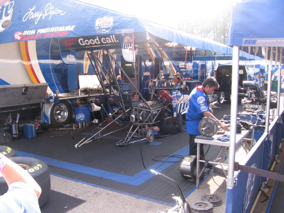 Racing team pit area with dragster chassis, tools, and mechanics working under a blue tent