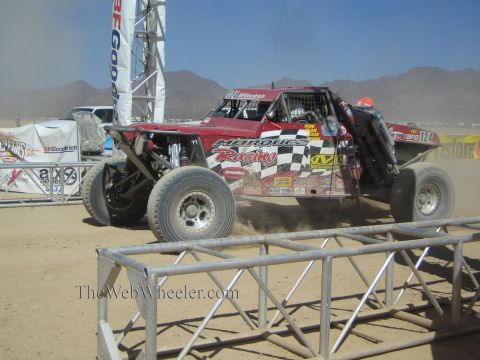 1990 Ford Bronco - red off-road race buggy numbered 124 with Marque Racing livery and checkered-flag graphics races at a desert start.